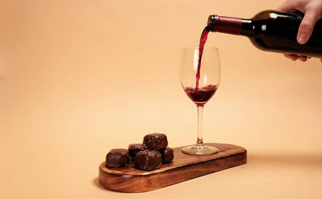 Red wine being poured into a wine glass beside milk chocolates on a wooden serving board for a chocolate and wine pairing.
