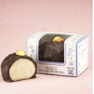 16-oz Dark Chocolate Vanilla Butter Cream Eggs with edible yellow decorative flowers on top. One (1) Egg cut in half to reveal white inside. Other egg whole in package with clear window to show outside of egg. Pictured on light purple backdrop.