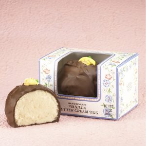 16-oz Milk Chocolate Vanilla Butter Cream Eggs with edible yellow decorative flowers on top. One (1) Egg cut in half to reveal white inside. Other egg whole in package with clear window to show outside of egg. Pictured on light purple backdrop.