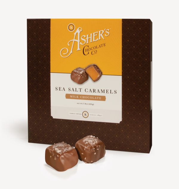 Milk Chocolate Sea Salt Caramels Traditional Box with yellow and white Asher's Chocolate Co. Label. Two (2) caramels on the front of the box reveal, color, size, shape, and texture of candies.Two (2) Dark Chocolate Sea Salt Caramels sit outside of the box to reveal size, color, and chocolate texture. Displayed on white background.