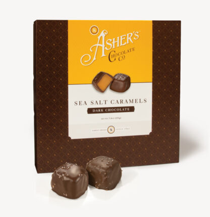 Dark Chocolate Sea Salt Caramels Traditional Box with yellow and white Asher's Chocolate Co. Label. Two (2) caramels on the front of the box reveal, color, size, shape, and texture of candies.Two (2) Dark Chocolate Sea Salt Caramels sit outside of the box to reveal size, color, and chocolate texture. Displayed on white background.