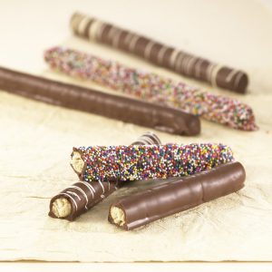 Assortment Pretzel Rods shown on tan background. Three (3) rods are shown in front, cracked in half. One (1) plain chocolate coated, one (1) with white chocolate drizzle, and one (1) with rainbow seeds. Three (3) whole pretzel rods are shown in the background.