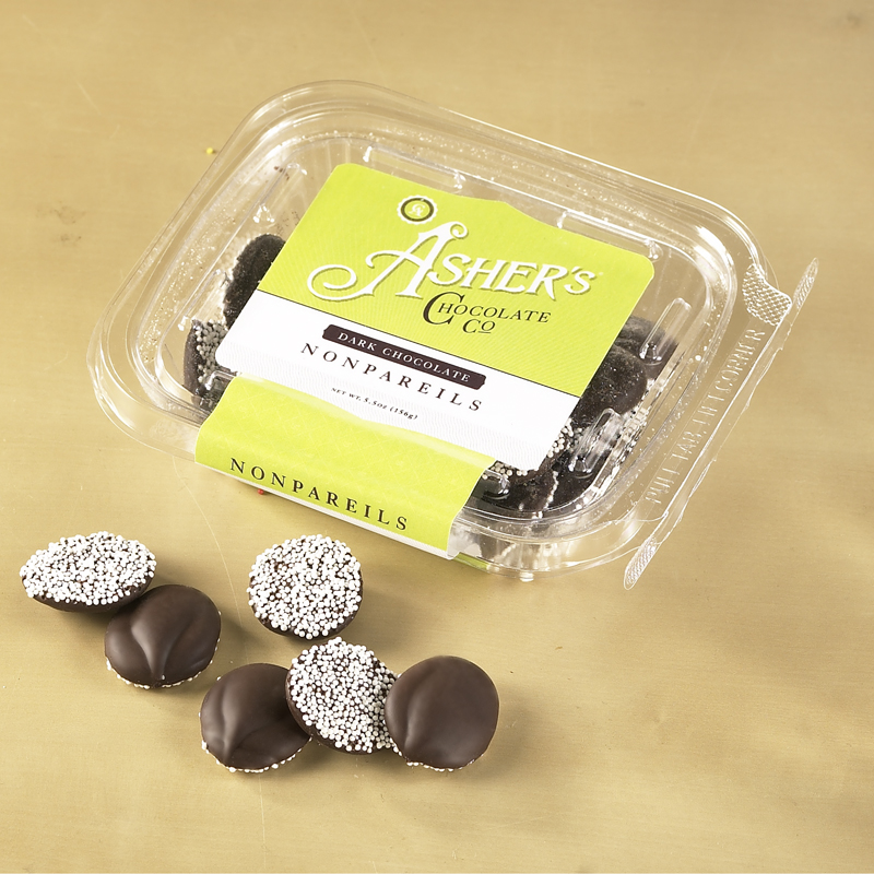 Dark Chocolate Nonpareils Packaged Fresh Pack with yellow and white Asher's label. Visible inside clear package are dark chocolate nonpareils with white seeds. Scattered outside package are whole dark chocolate nonpareils with white seeds.