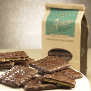 Milk Chocolate Graham Crackers Coffee Bag in background with broken and whole Grahams on plate in front. Coffee Bag is brown with teal details. Clear plastic circular window on front displays Milk Chocolate Grahams inside.