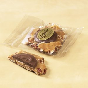 Hand Crafted Milk Chocolate Caramel Praline shown in cello bag with gold Asher's sticker. One (1) praline is shown outside of the bag, cut in half, to reveal the chocolate topping, nuts, and caramel filling.
