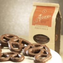 Milk Chocolate Pretzel Coffee Bag in background with whole three (3) ring pretzels on white plate in front. Coffee Bag is brown with orange details. Clear plastic circular window on front displays Milk Chocolate Pretzels inside.