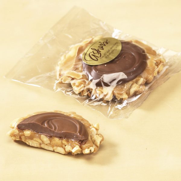 Cashew Praline Hand Crafted Milk Chocolate shown in cello bag with gold Asher's sticker. One (1) praline is shown outside of the bag, cut in half, to reveal the chocolate topping, nuts, and caramel filling.