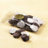 Dark Chocolate Nonpareils clear cello bag.Dark chocolate drops are scattered outside of clear cello bag to reveal size, color, and texture. White seeds coat one side of the drop. Pictured on tan background.