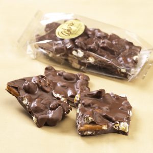 Milk Chocolate Boardwalk Crunch in clear Cello Bag with gold Asher's Sticker. Broken chunks of crunch lie outside of cello bag to reveal the chocolate toppings.