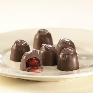 Milk Chocolate Cordial Cherries sit on white plate. One (1) cordial is cut open to reveal a sweet, red cherry inside.