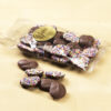 Milk Chocolate Nonpareils clear cello bag. Milk chocolate drops are scattered outside of clear cello bag to reveal size, color, and texture. Rainbow seeds coat one side of the drop. Pictured on tan background.
