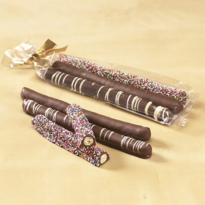 Assortment Pretzel Rods in Cello Bag show three (3) chocolate covered pretzel rods in a clear cello bag with gold bow. Three (3) rods are shown outside the bag, one (1) plain chocolate coated, one (1) with white chocolate drizzle, and one (1) with rainbow seeds. Rainbow seed pretzel is snapped in half to reveal the pretzel inside.