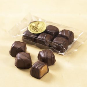Vanilla Caramels 4-oz Cello Bag with six (6) candies inside a clear cello bag with Asher's gold sticker on the front. Four (4) chocolates are shown outside the bag, one (1) being open to reveal the vanilla caramel center. Rest are shown whole with dark chocolate drizzle.