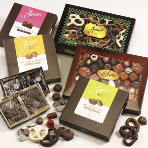 Various Boxed Chocolates shown on white background. Two (2) boxes of nonpareils with rainbow and white seeds are shown in a box with a thin gold ridden. Cherry Cordials, Sea Salt Caramels, and Pecan Patties are shown in boxes. Larger boxes of assorted chocolates and three (3) ring chocolate covered pretzels are shown. Scattered around the boxes are loose pieces of chocolate to reveal colors, sizes, and textures.