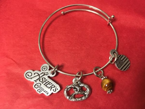 Asher's Chocolate Co. Silver Charm Bracelet shown on red background. Silver Charms include an Asher's Chocolate Co. Logo, a three (3) ring pretzel twist, a brown stone and a circular American flag.