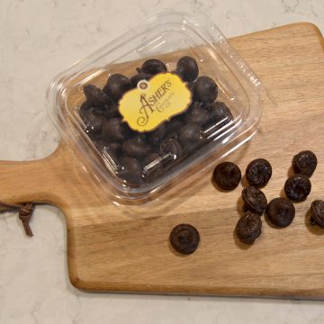 Dark Chocolate Wilbur Bubs Fresh Pack displayed on a wooden board. Some dark chocolate bubs are scattered outside of fresh pack, displaying shape, and color. Rest of bubs fill fresh pack with gold and brown Asher's sticker labeling the lid.