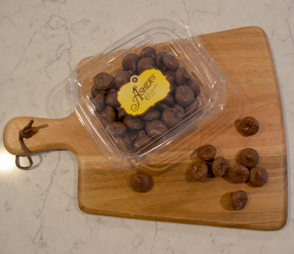 Milk Chocolate Wilbur Bubs Fresh Pack displayed on a wooden board. Some milk chocolate bubs are scattered outside of fresh pack, displaying shape, and color. Rest of bubs fill fresh pack with gold and brown Asher's sticker labeling the lid.