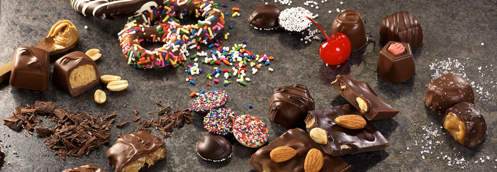 Ingredient Slider of Chocolate Assortments. Chocolate shavings, peanuts, and a wooden spoon of peanut butter lie near milk chocolate smoothies. Three (3) ring gourmet pretzel with rainbow sprinkles lies in a pile of rainbow sprinkles. Nonpareils are shown with white seeds and rainbow seeds. Cheer cordials are shown with a red cherry. Milk and dark Sea Salt Caramels are shown surrounded by course sea salt. Almond bark is broken with whole almonds shown on top.