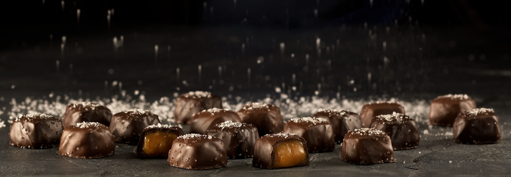 An assortment of milk and dark chocolate Sea Salt Caramels lie on a dark background covered in course salt that crystallizes in the background.