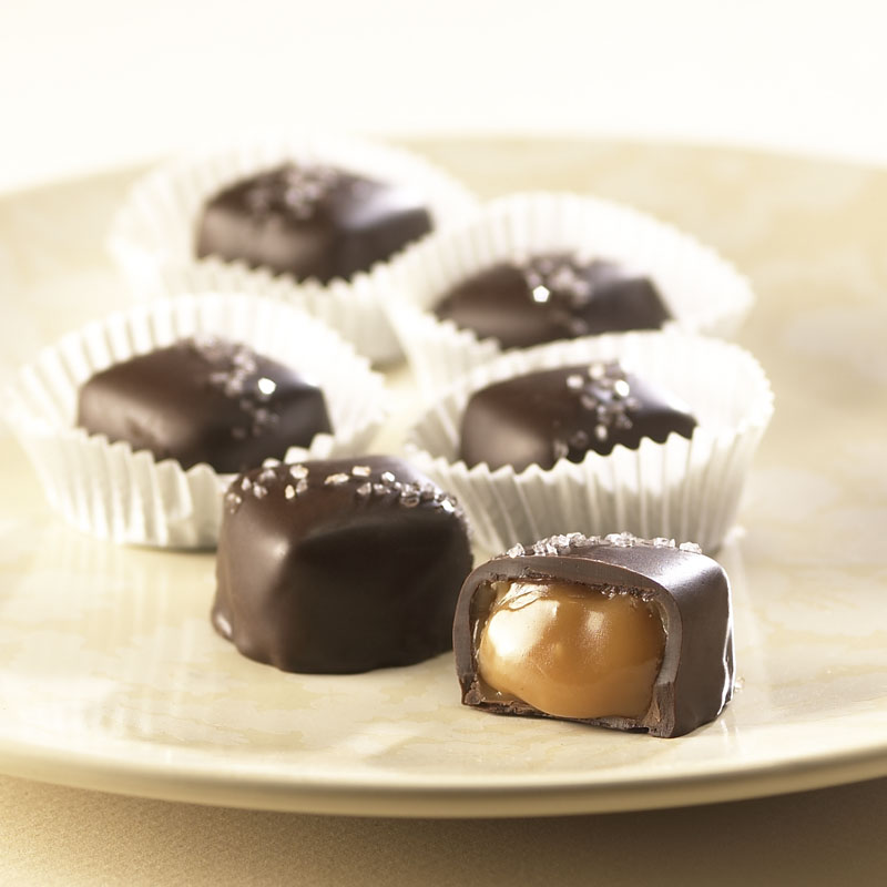 The Asher's Chocolate Co. sweet & salty snacks, Sea Salt Caramels laid out on a tan plate with one cut open to show the gooey caramel inside.