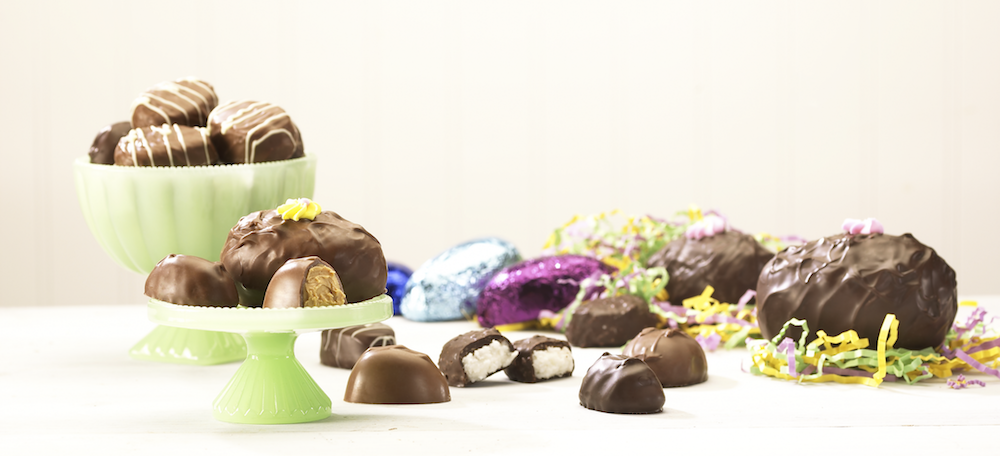Large Easter Egg with yellow flower sits with two (2) smaller milk chocolate peanut butter eggs, one of which is cut open to reveal smooth brown center, atop a green cake display. Next to the display lies four eggs, one of which is dark chocolate coconut. In the background there are three (3) wrapped eggs in purple, light blue, and dark blue foil. There is a large green bowl with milk chocolate eggs that have white drizzle. In the background, two (2) large eggs covered in chocolate with pink flowers sit on yellow, green, and purple hay.