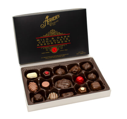 Open box of Gourmet Chocolate Assortment 8oz., a selection of gourmet chocolates in an array of shapes and styles—some decorated with drizzles or nuts—arranged on a white background. The black lid features sophisticated gold and red lettering.