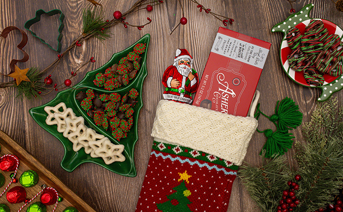 Flat-lay of old-fashioned Christmas candies arranged on a rustic wooden table, featuring Asher’s chocolate bars, a foil-wrapped Santa chocolate, festive nonpareils, white chocolate-covered treats in a green ceramic Christmas-tree dish, and a holiday stocking filled with chocolates. Surrounding decorations include cookie cutters, greenery, berries, and a plate of striped peppermint-style candies.