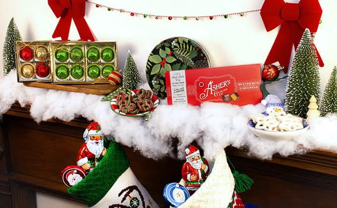 Festive holiday display featuring Asher’s chocolates arranged on a mantel decorated with faux snow, miniature Christmas trees, and red ribbon bows. Gift-ready treats include boxed chocolate assortments, a red Asher’s chocolate bar, foil-wrapped Santa chocolates, white chocolate-covered pretzels in a bowl, and a plate of striped holiday candies, with filled Christmas stockings hanging below.