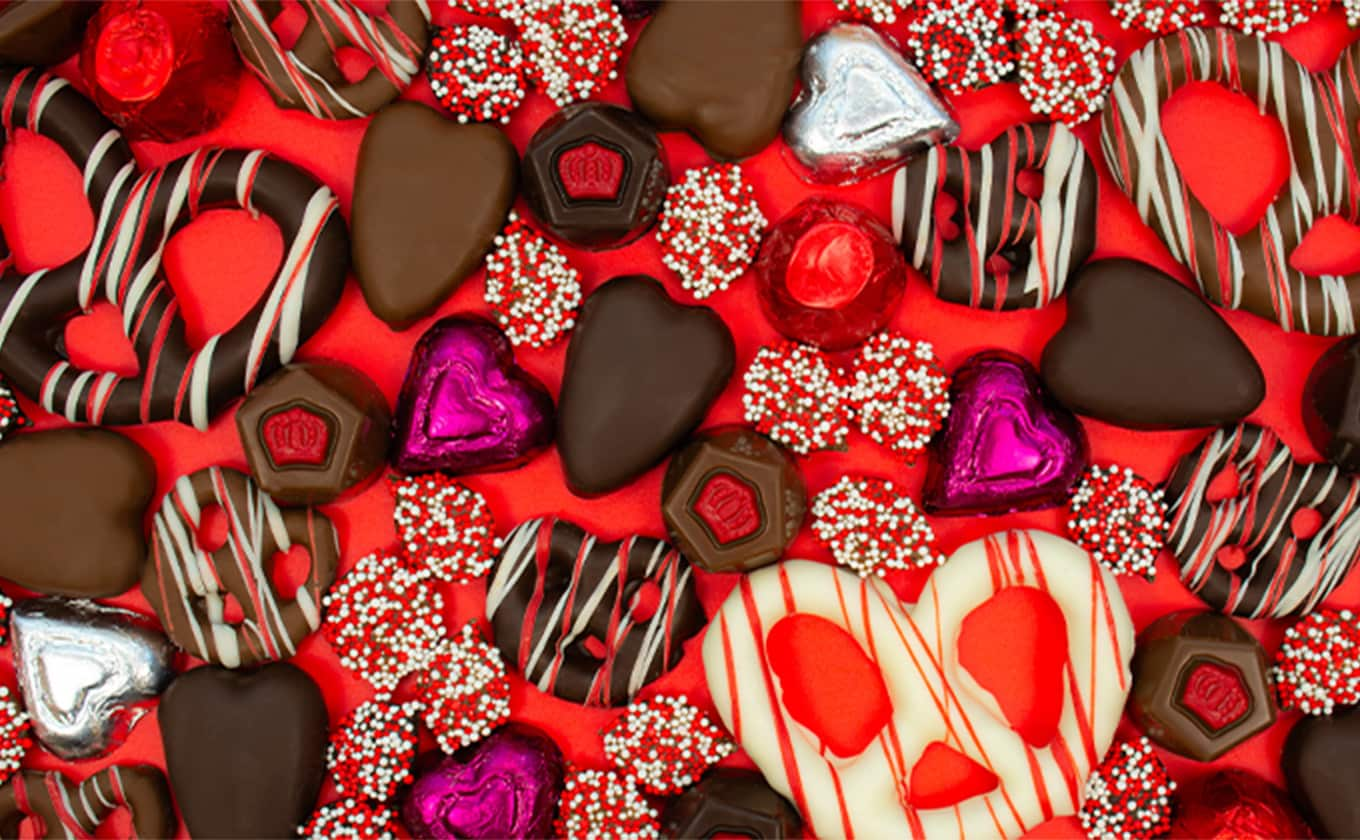 Close-up assortment of Valentine’s Day chocolates featuring heart-shaped milk and dark chocolates, decorated truffles, foil-wrapped hearts, and red and white chocolate drizzles on a bright red background