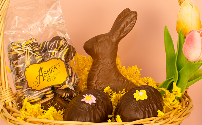 Easter basket filled with shredded yellow paper grass, a large milk chocolate bunny, two chocolate eggs, and a bag of chocolate covered pretzels, with spring tulips in the background.