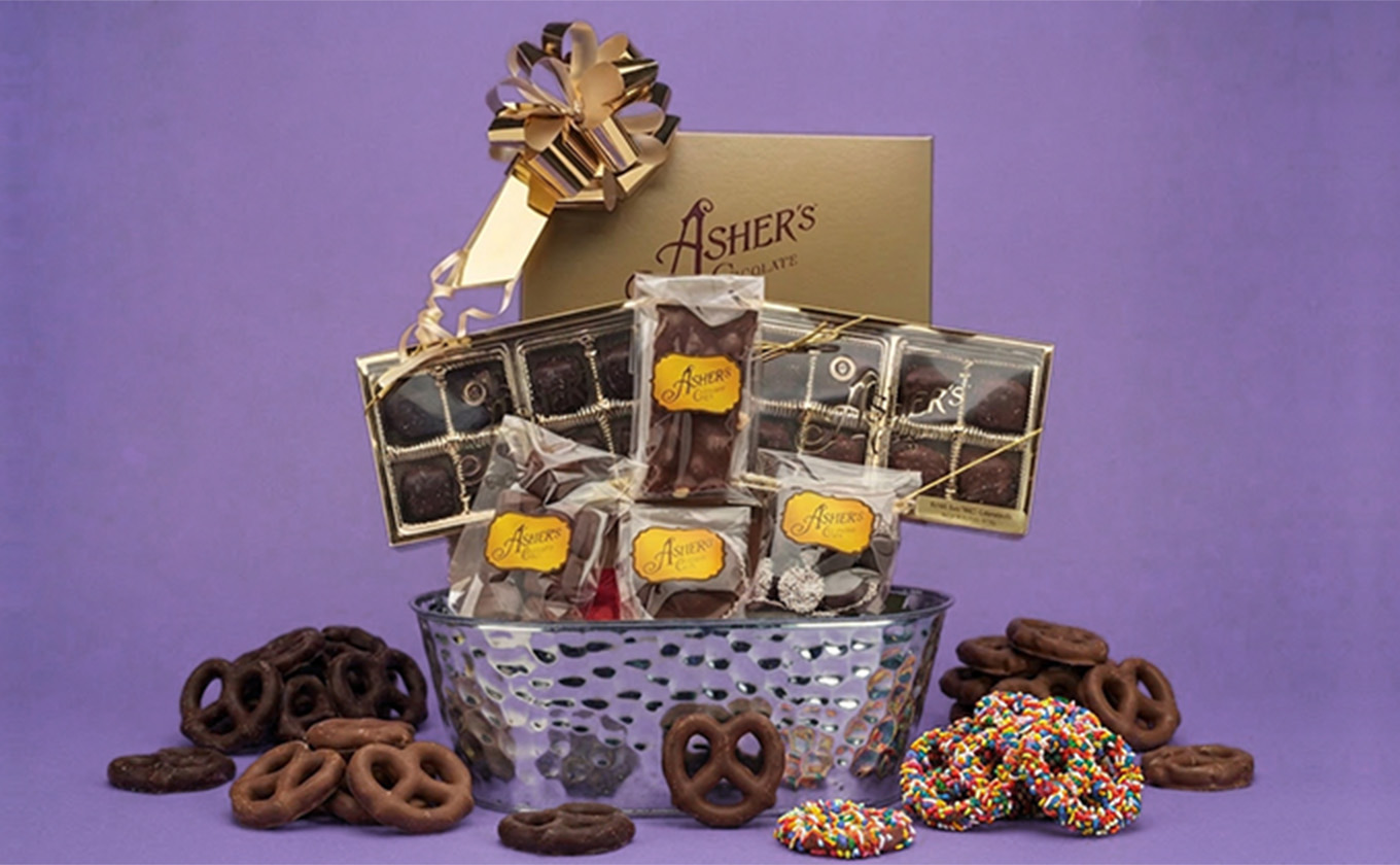 Chocolate gift basket from Asher’s Chocolate Co. featuring assorted chocolate-covered pretzels, cookies, and confections, arranged with a gold bow for a Mother’s Day gift idea