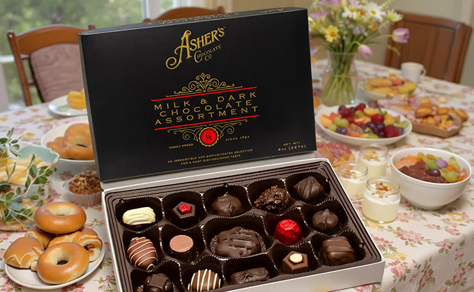 A box of Asher's assorted gourmet chocolates on a table decorated for Mother's Day with a floral tablecloth and spread of bagels and other brunch foods.