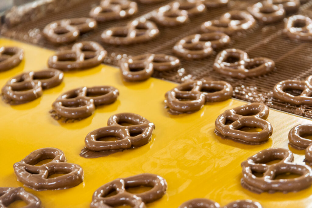 Chocolate-covered pretzels move along a yellow conveyor belt in a factory, reflecting Asher’s chocolate history, with some pretzels coated in rich chocolate and others still being processed on a wire rack.