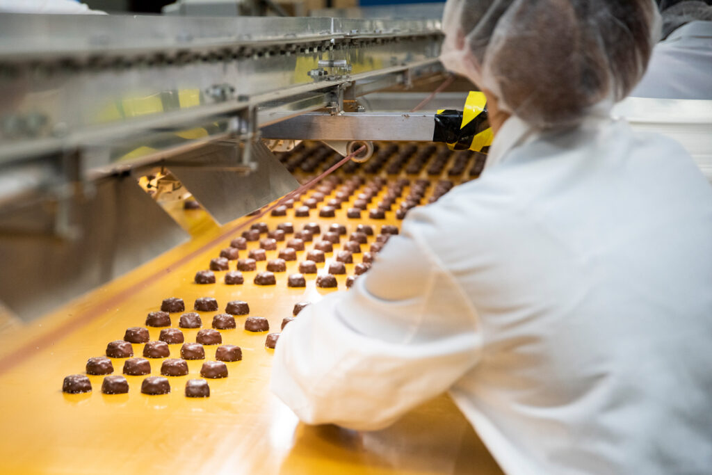A worker in a white lab coat and hairnet oversees chocolates moving along a conveyor belt in a factory setting, embodying the meticulous standards that have shaped Asher’s chocolate history.