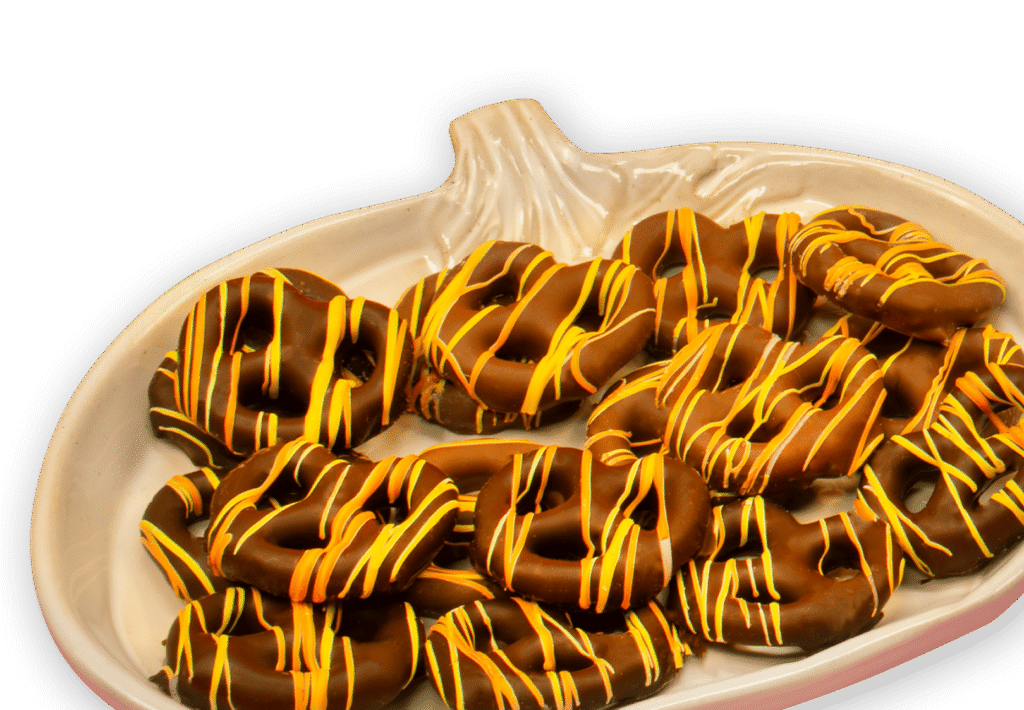 A white platter shaped like a pumpkin holds chocolate-covered pretzels drizzled with orange and yellow icing, arranged in a pile against a white background.