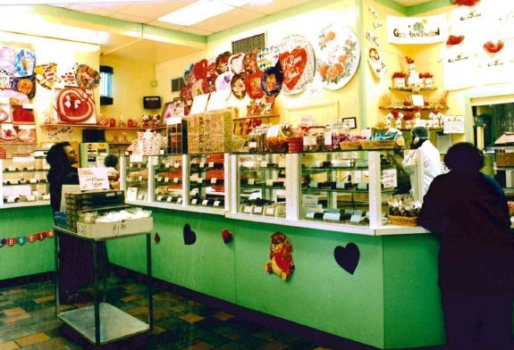 A candy shop decorated for Valentine’s Day, with heart-shaped boxes and gifts on display, customers at the counter, and shelves filled with assorted treats behind glass—echoing Asher’s chocolate history in every detail.