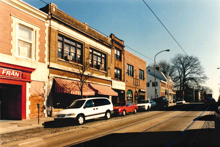 A sunny street scene with parked cars, brick storefronts, a red awning, and leafless trees captures the charm of a vibrant neighborhood. Power lines and a streetlamp run along the sidewalk, while the clear blue sky offers perfect weather for exploring local shops—perhaps even learning about Asher’s chocolate history at a nearby confectionery.