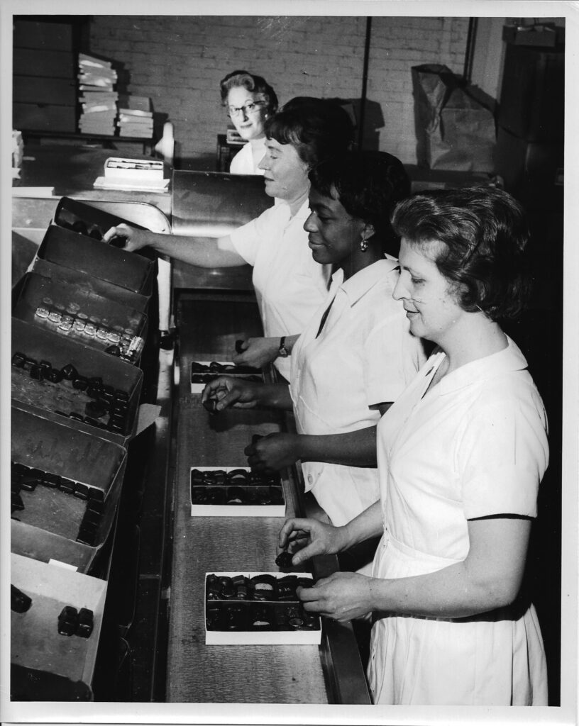 Four women in white uniforms work at a factory assembly line, placing chocolates into boxes. Reflecting the rich Asher’s chocolate history, they are standing in a row, focused on arranging the chocolates neatly inside each box.