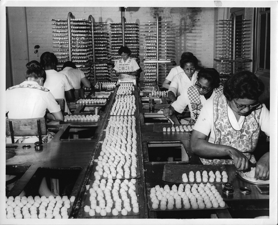 Black-and-white photo of women working at a long table, packaging rows of marshmallow peep candies on trays in a factory setting—a glimpse into asher’s chocolate history. Shelves with more trays appear in the background.