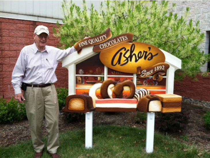 An older man in a cap stands next to a colorful Ashers Chocolates bench, decorated with large chocolate sculptures that celebrate Asher’s chocolate history, outside in front of a building and some green bushes.