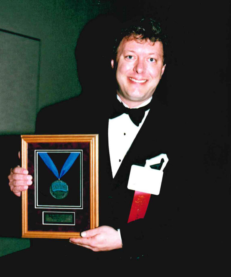 A person in a tuxedo smiles while holding a framed award with a blue ribbon medal, celebrating their achievements connected to Asher’s chocolate history. A name badge and red ribbon are pinned to the jacket. The background is dark.