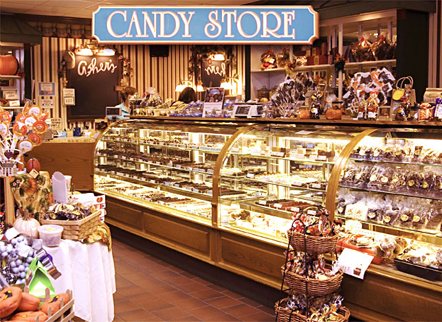 A brightly lit candy store with glass display cases filled with assorted candies and treats, baskets of sweets on the floor, and a blue CANDY STORE sign above the counter—the perfect place to experience a taste of Asher’s chocolate history woven into every delightful bite.