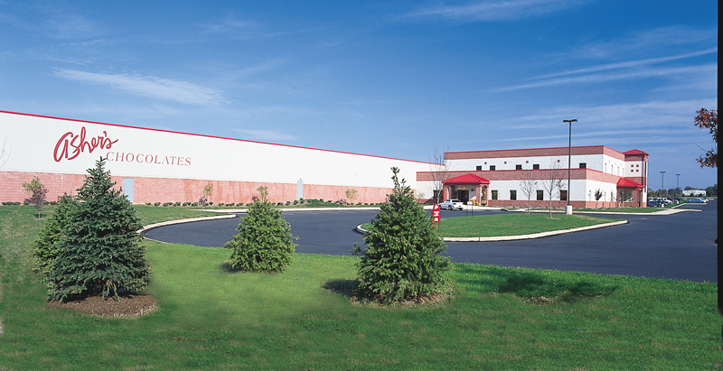 The large building labeled Ashers Chocolates, with its red roof entrance and neatly landscaped lawn dotted with small evergreen trees, stands as a testament to Asher’s chocolate history. A paved parking lot stretches out beneath the clear blue sky, welcoming visitors to this iconic location.