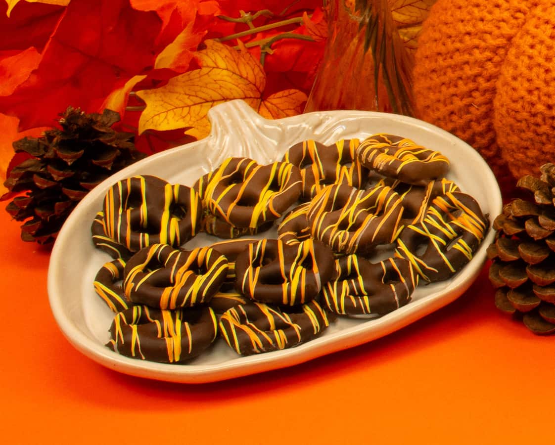 Chocolate-covered cookies with orange and yellow drizzle arranged on a pumpkin-shaped plate. Autumn leaves, pinecones, and a knitted pumpkin decorate the orange background, giving a fall or Thanksgiving theme.