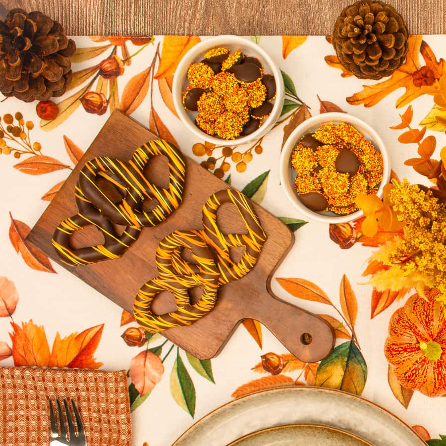 Wooden board with chocolate-covered pretzels drizzled with yellow icing on a fall-themed table, surrounded by bowls of chocolate candies, pinecones, and autumn flowers.