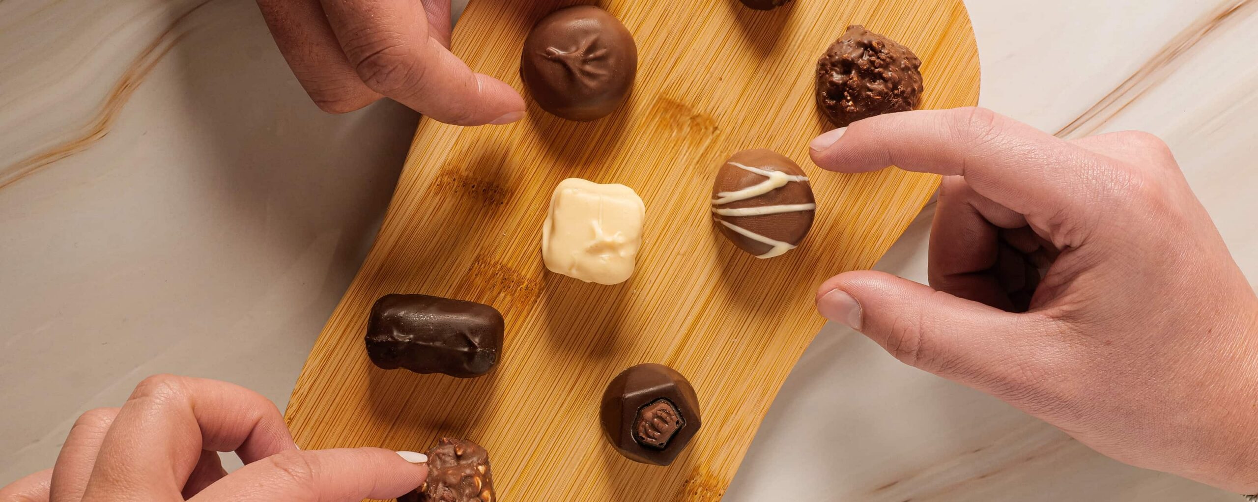 Hands reaching for a variety of assorted chocolates arranged on a wooden board.