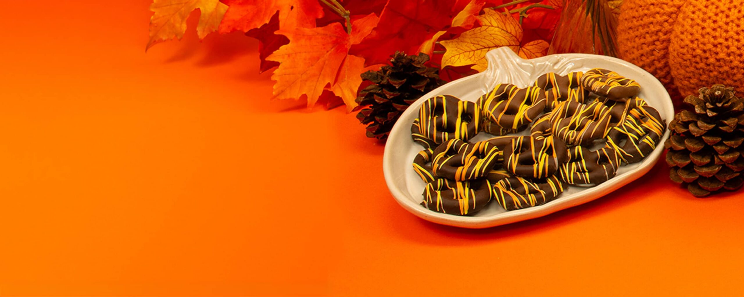 Chocolate-covered pretzels with yellow icing drizzle on a white pumpkin-shaped plate, surrounded by autumn leaves and pinecones on an orange background.